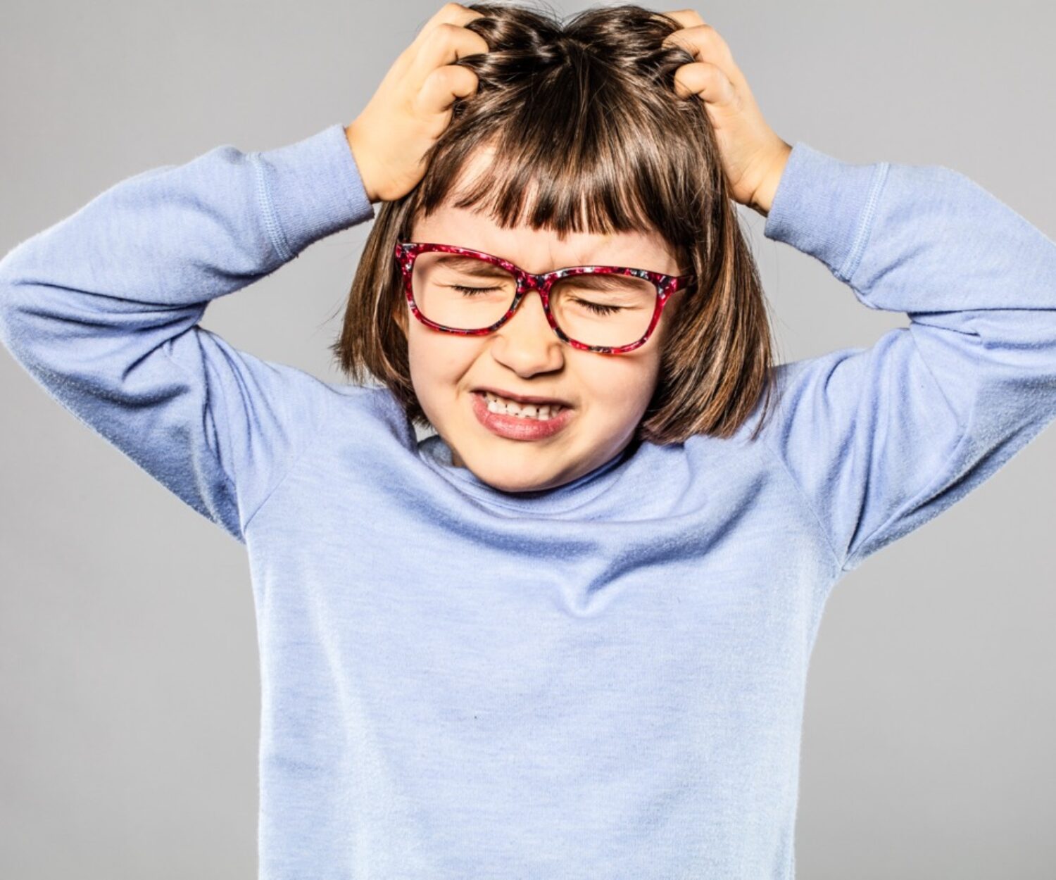irritated 6-year old young girl with eyeglasses pulling out her hair for itchy allergies or lice or scratching her head for nervous disagreement, grey background