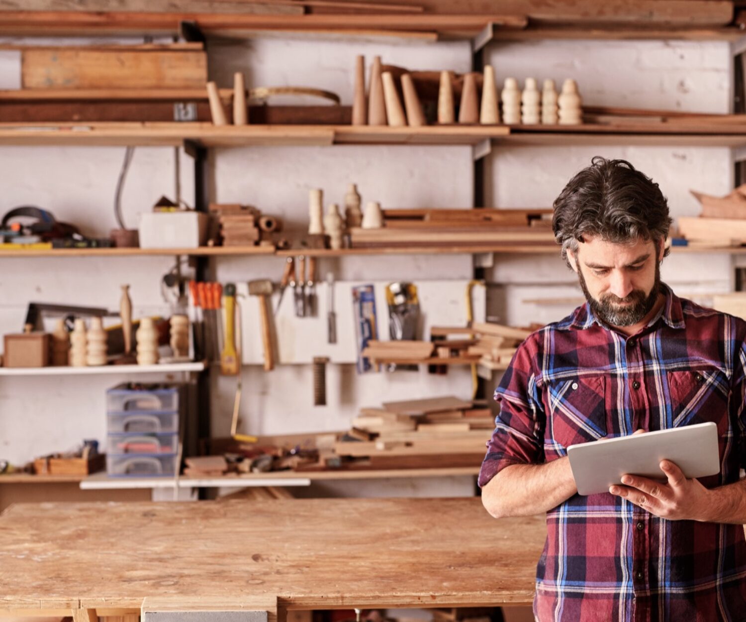 Artisan woodwork studio with shelving holding pieces of wood, with a carpenter standing in his workshop using a digital tablet